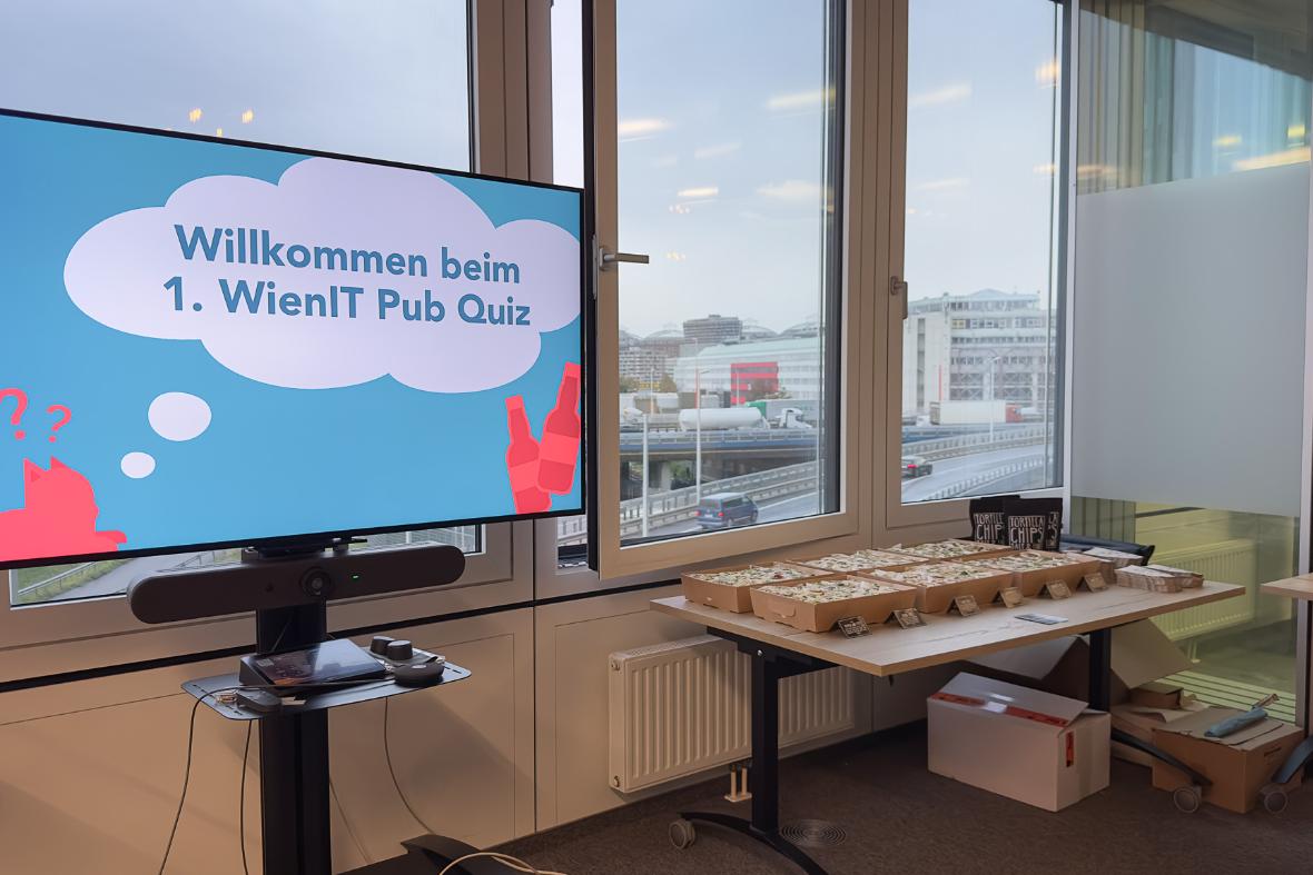 Das Bild zeigt einen Bildschirm, auf dem "Willkommen zum WienIT Pub Quiz" steht - im Hintergrund ist das Buffet mit Essen zu sehen. 