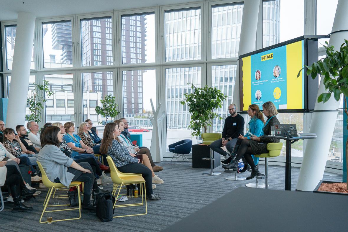 Mehrere Menschen sitzten auf gelben Sesseln. Die 3 Podiums-Gäste sitzen auf Bar-Hockern, im Hintergrund sieht man ein großen Bildschirm.